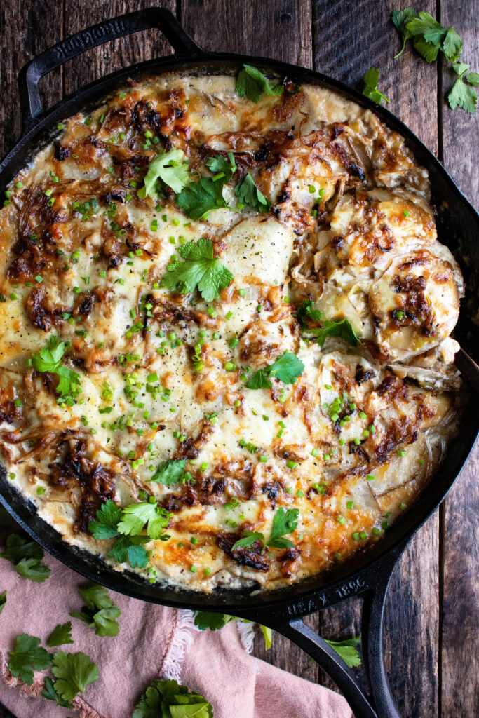 a skillet of scalloped potatoes out of the oven with melted cheddar and herbs over top