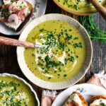bowls of Asparagus Soup topped with a drizzle of cream and fresh herbs with prosciutto goat cheese toast on the side