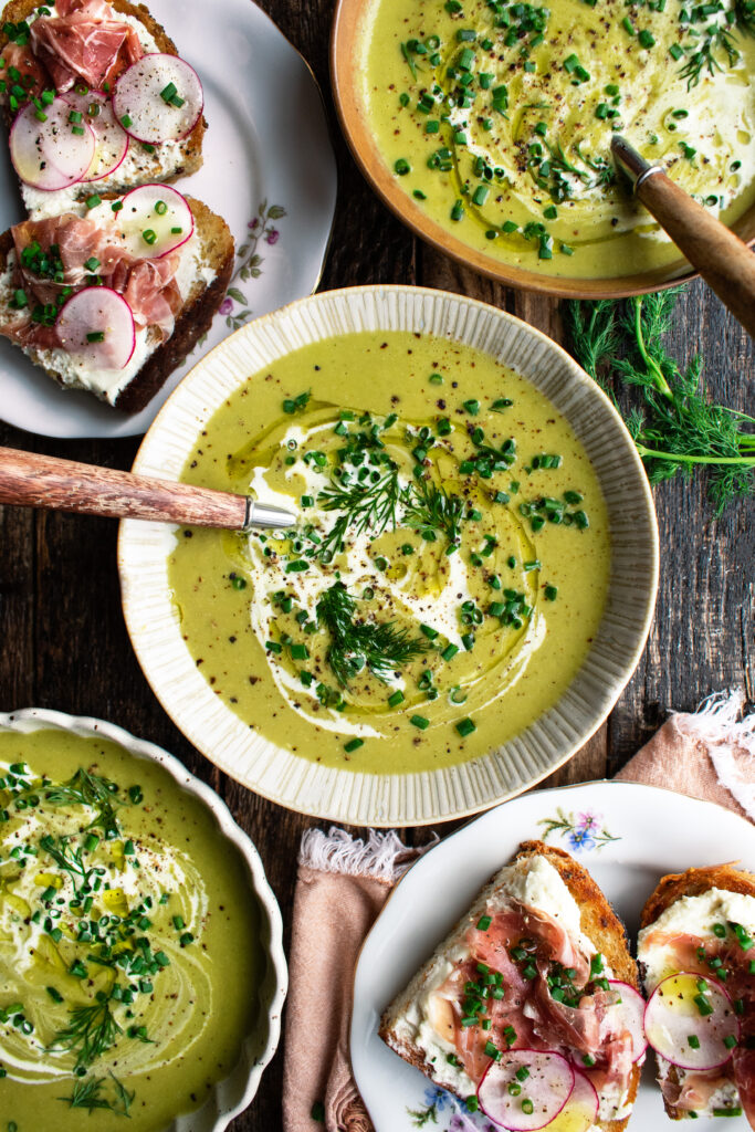 bowls of Asparagus Soup topped with a drizzle of cream and fresh herbs with prosciutto goat cheese toast on the side