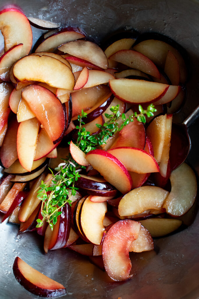 sliced plums and fresh thyme macerating in sugar in a mixing bowl