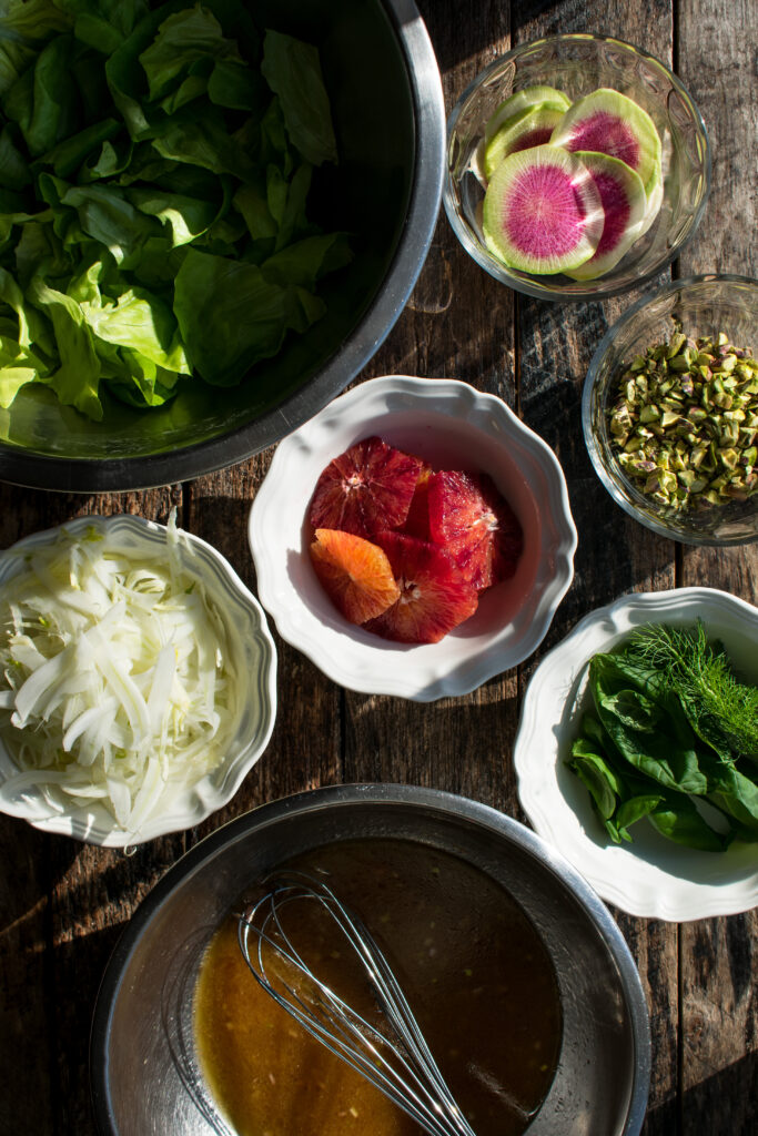 ingredients for the salad prepped in bowls: butter lettuce, watermelon radishes, fennel, blood oranges, pistachios, basil, and spicy citrus vinaigrette
