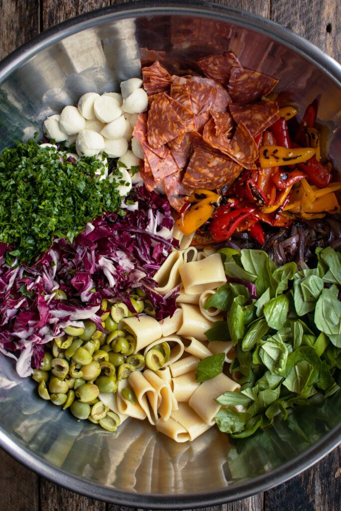 pasta salad ingredients in a bowl: basil, pasta, red onion, caramelized peppers, salami, fresh mozzarella, parsley, radicchio, and olives